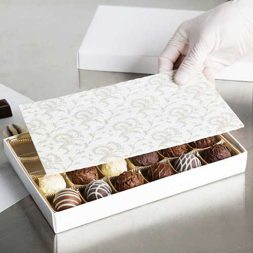 A gloved hand lays a protective packing layer over a box full of chocolates