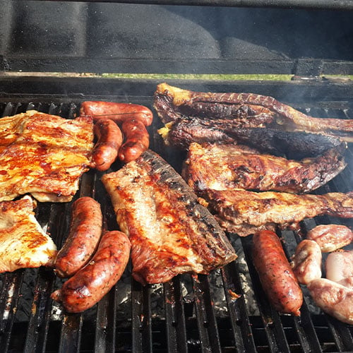 An assortment of asado meats grilling including chorizo, ribs, chicken breasts, and steak