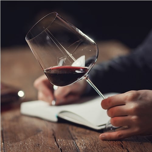 a person's hand grips a wineglass by its stem to swirl it on a wooden table