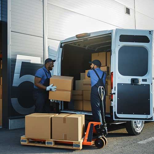 two workers talk while using a hand pallet truck to load a delivery truck with cardboard boxes