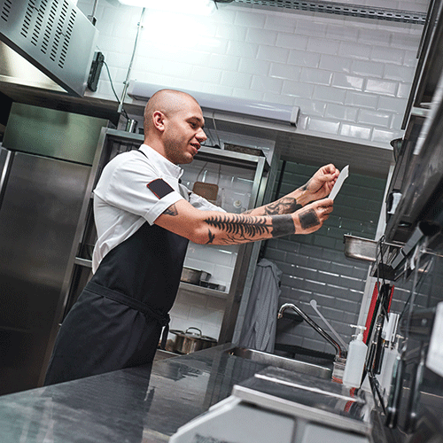 bald male chef with tattoos on his arms looking at order list in a restaurant kitchen