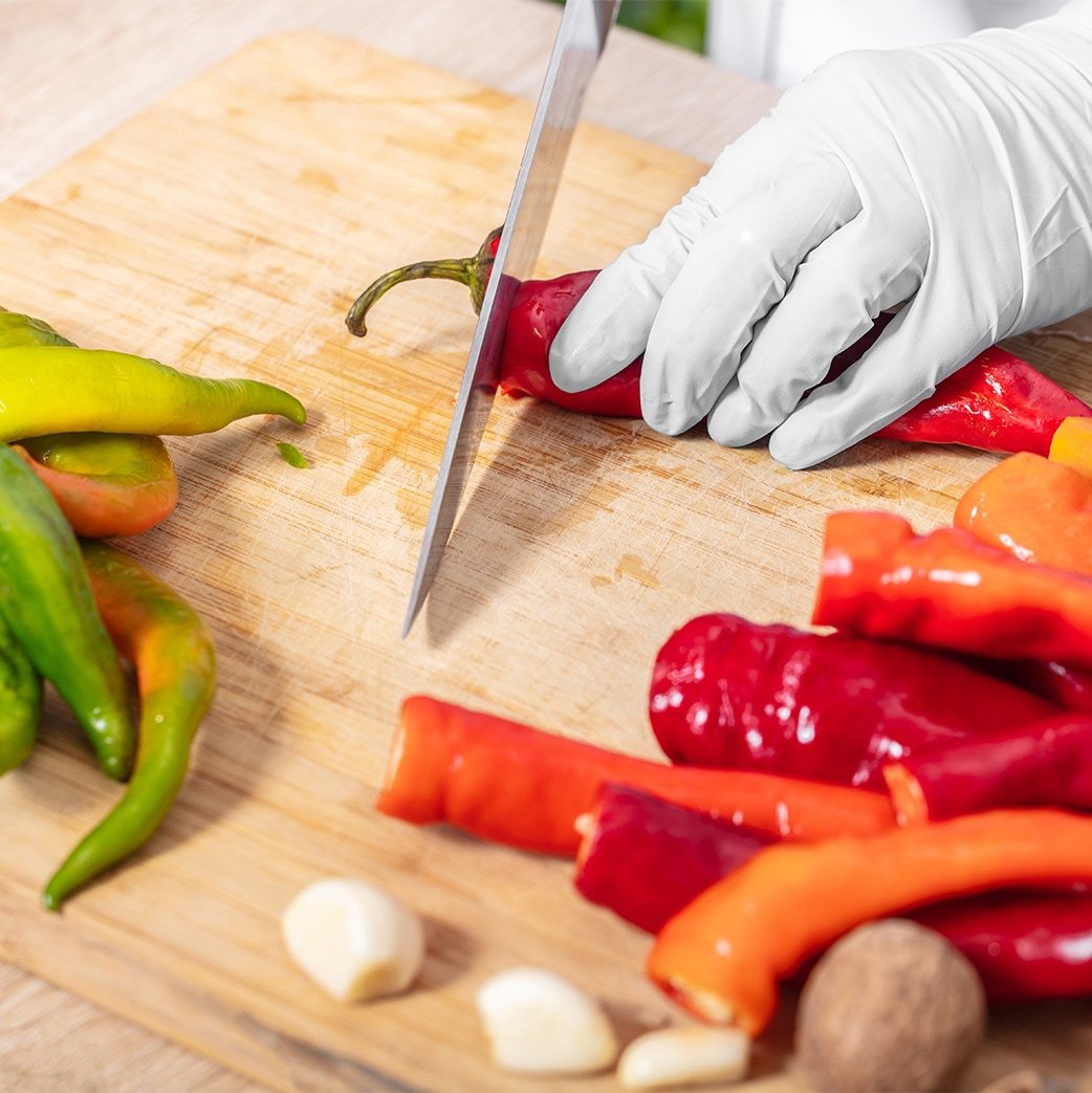 gloved hand holding a red chili on a cutting board and slicing the top with a chef knife