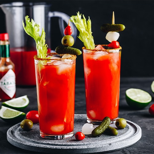 Two bloody mary drinks on a gray background with ingredients surrounding