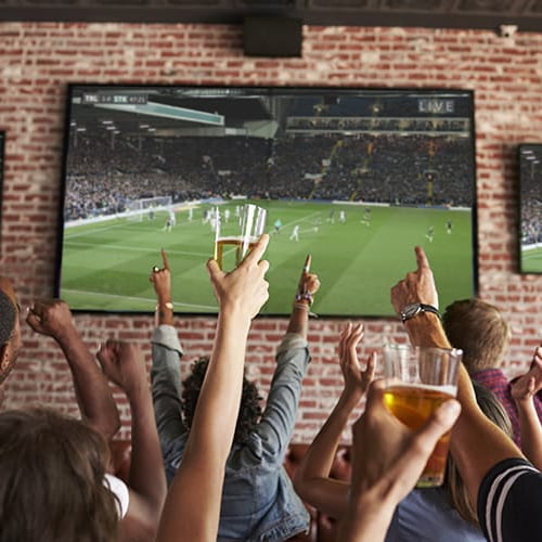 People raising glasses and hands in front of football game on screen