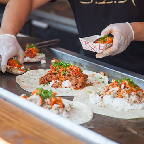 white gloved hands assembling a line of tortillas topped with rice, beef, orange sauce