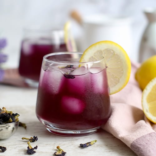 A mocktail containing iced butterfly pea flower tea, juice, and a sliced lemon