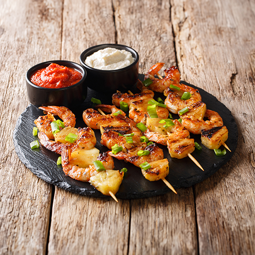 grilled shrimp and pineapple skewers on a black slate plate with cocktail and tartar sauce in black ramekins