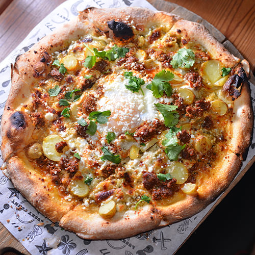 California pizza on wooden table with tomatoes, garlic, mushroom, and pepper next to it 