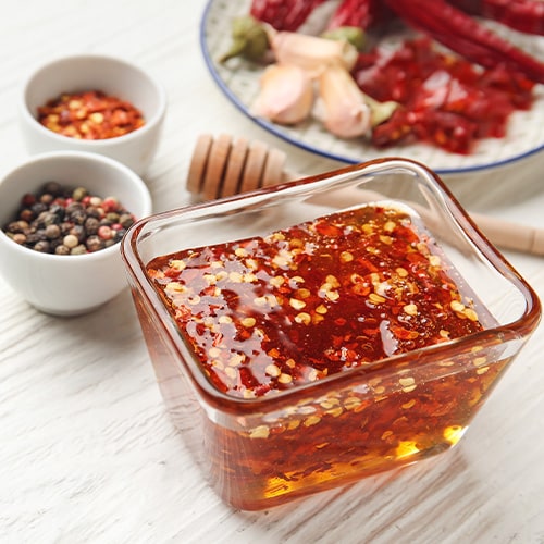 a bowl with hot honey with bowls of red pepper flakes and peppercorns beside it on a light wooden background