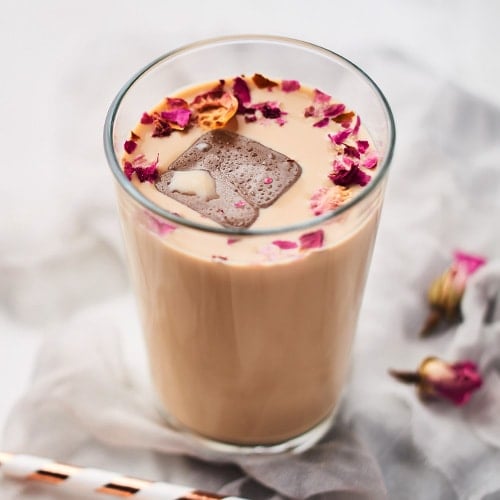 iced coffee with rose and cardamom in a tall glass surrounded by dried roses and straws on white and gray silk background