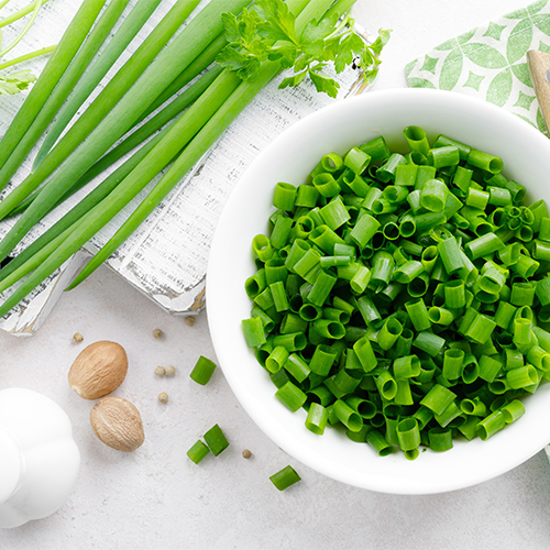 An overhead view of a white table with a large bowl of cut green onions on top next to a bundle of green onions