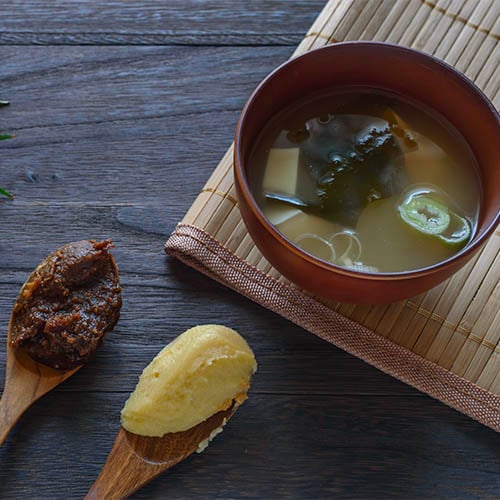 two types of miso on wooden spoons in front of a bowl of miso soup