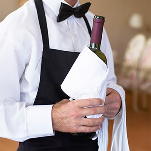 A waiter dressed in a white shirt, black bow tie, and black apron holds a bottle of wine wrapped in a white cloth.