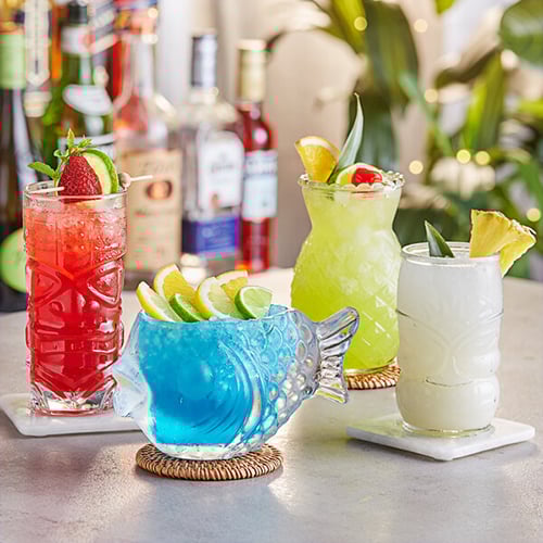 four colorful mixed drinks in different-shaped tiki glassware with fruit garnishes
