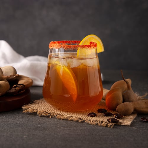 stemless glass filled with a tamarind-infused liquid, red salted rim, and lemon wedges