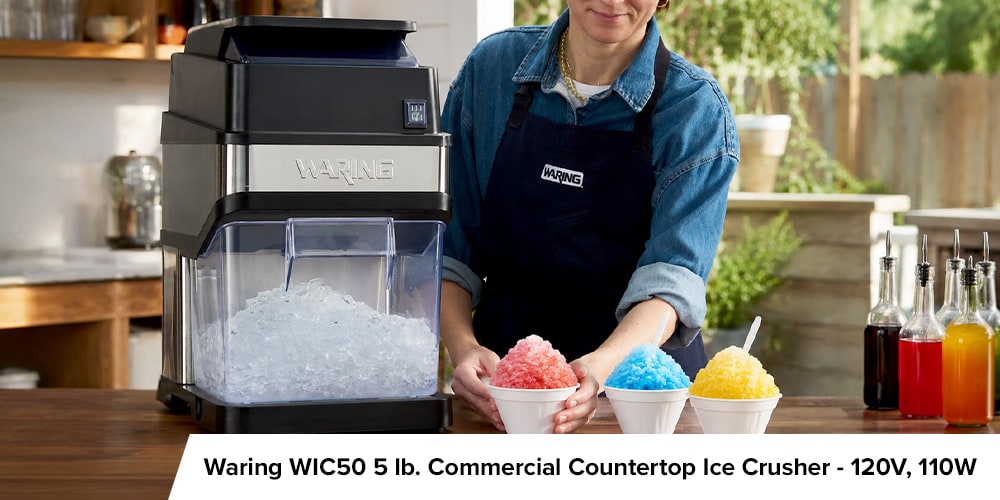 Waring commercial countertop ice crusher beside a server presenting red, blue, and yellow snow cones.