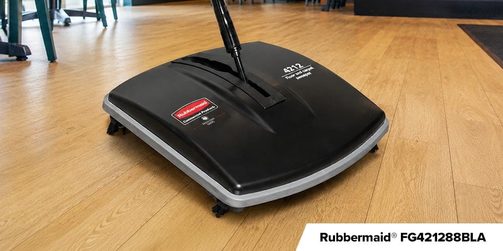 Rubbermaid FG421288BLA - Close-up of a black Rubbermaid commercial floor sweeper positioned on a light wood floor.