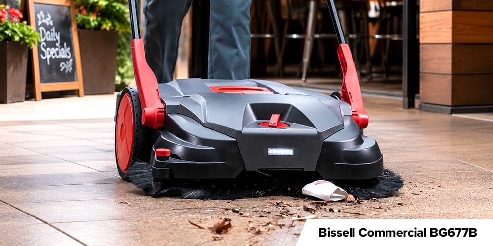 Bissell Commercial BG677B - Person pushing a large black-and-red Bissell commercial sweeper over debris outside a restaurant entrance.