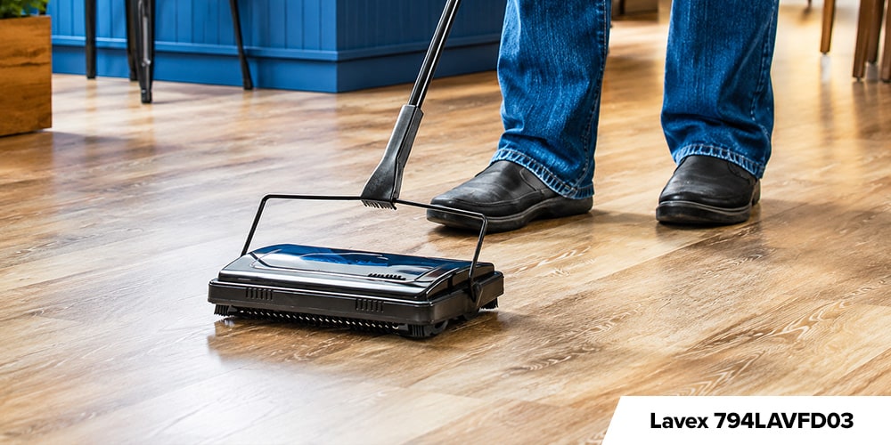 Lavex 794LAVFD03 - Person using a black manual floor sweeper on a wood floor in a brightly lit café or restaurant.
