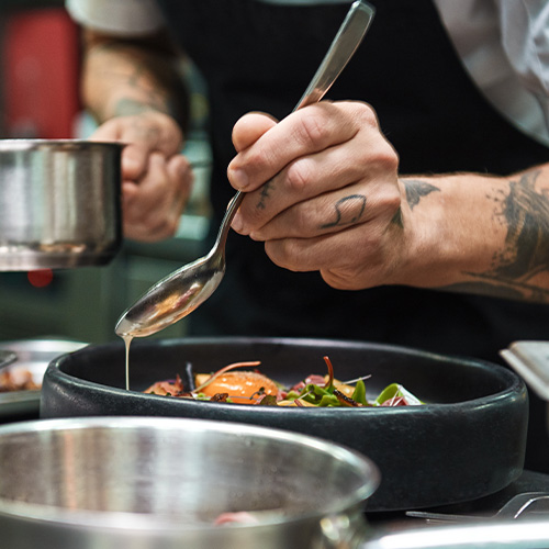 Close up photo of chef hands with several tattoos adding a sauce to italian pasta Carbonara