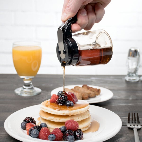 A person using a Tablecraft Modern Glass Syrup Dispenser with black ABS top to pour syrup over a stack of pancakes with berries.