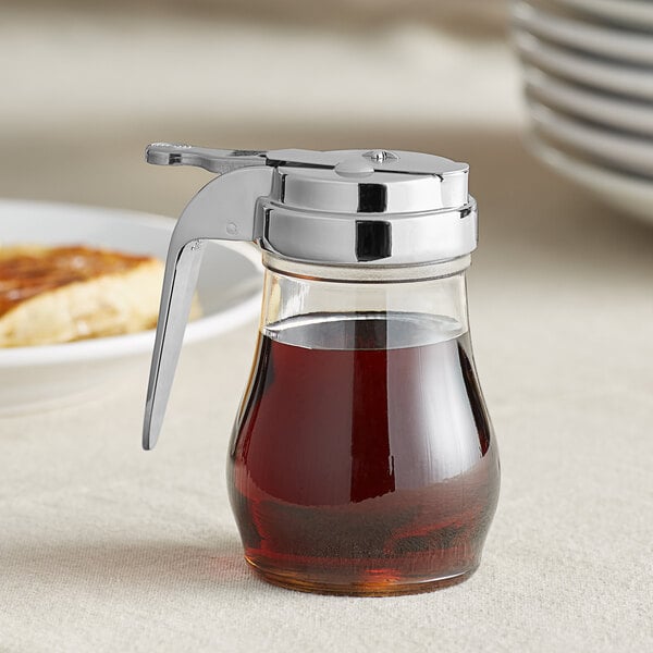 A clear polycarbonate syrup server with a chrome top and brown syrup inside.