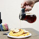 A hand using a Tablecraft glass teardrop syrup dispenser to pour syrup on a plate of waffles with bananas and chocolate chips.